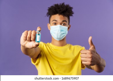 Close-up Portrait Of Upbeat Young Hispanic Man In Medical Mask Holding Ampoule With Covid19 Vaccine, Coronavirus Drug, Show Thumbs-up, Getting Shot At Ambulance, Standing Purple Background