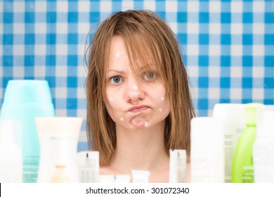 Close-up portrait of upset cute girl without makeup, with white spots of cosmetic on her problem face skin, standing in bathroom surrounded by various cosmetic products. Skincare and beauty concept