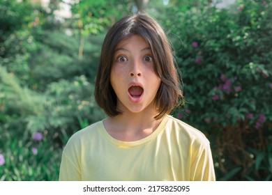 Close-up portrait of a very surprised teenage girl with wide open eyes and mouth against the background of greenery.