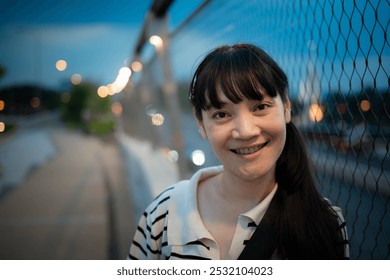 A close-up portrait of a woman against a chain-link fence, her smile glowing under the evening lights. The blurred city lights create a soft bokeh effect in the background. - Powered by Shutterstock