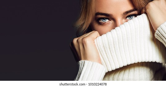 A close-up portrait of a woman. A beautiful young girl with blue eyes covers her mouth with a collar of white sweaters.fashion, beauty, makeup, cosmetics, beauty salon, style, personal care, posture.