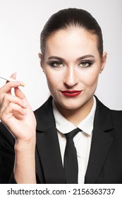 Close-up Portrait Of Woman With Red Lipstick Holding Cigarette In Her Hands Near Face And Looking To Camera. Model With Conservative Hairstyle And Wearing Black Classic Suit With Tie And White Shirt