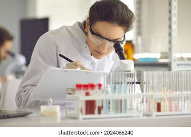 Closeup portrait of woman scientist working in lab. Female researcher sitting at desk looking through microscope at blood and chemical test sample and making notes in clipboard. Biochemistry work