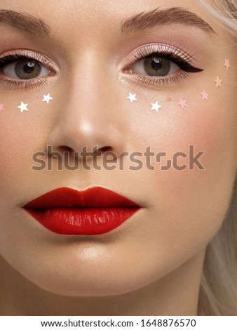 Close-up portrait of a woman with shiny clean skin and curly blond hair. Rhinestones stars on the eyes, evening makeup and red lipstick. soft care, full lips, long eyelashes and thick eyebrows