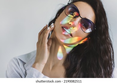 Closeup Portrait Of Woman In Trendy Glasses With Ray Of Rainbow Light On Her Face. Rainbow Optical Flare From The Window Projected On Her Face. Art Beauty Portrait. Girl With Rainbow Shining Face