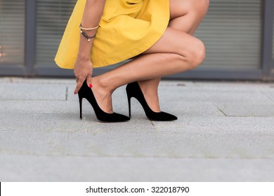 ハイヒールの女性の脚の接写。黄色いドレスを着た女性がオフィスビルの近くで右脚に座り、触っている。