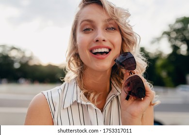 close-up portrait of young attractive stylish blonde woman in city street in summer fashion style dress wearing sunglasses, laughing with candid smile white teeth, sunny