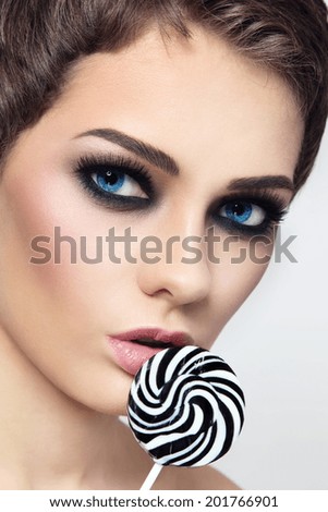 Close-up portrait of young beautiful blue-eyed brunette with smoky eyes and fancy striped lollipop in her hand