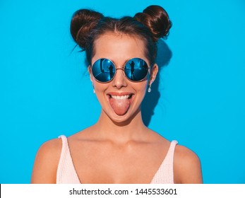 Closeup portrait of young beautiful sexy smiling hipster woman in sunglasses.Trendy positive girl going crazy.Model isolated on blue wall.Shows her tongue