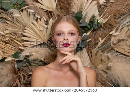 Closeup portrait of young beautiful woman with a healthy skin of the face. looks straight and hand fozle face Blonde girl with burgundy lips against a background of spring dry field flowers. 