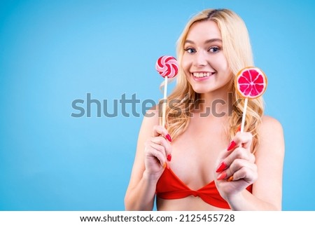 Closeup portrait of young blond woman in red swimsuit on blue background. Happy and joyful girl is holding bright colorful lollipop sweets candies in hands.  Vacation on sea beach concept. Summer mood