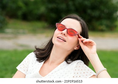 Closeup portrait young caucasian brunette woman in trendy sunglasses. Dreamy girl in white dress and modern red sunwear looking up at sky