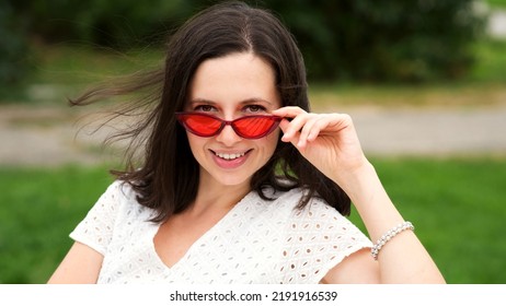 Closeup portrait young caucasian brunette woman in trendy sunglasses. Attractive girl in white dress and modern red sunwear looking over glasses at camera. 