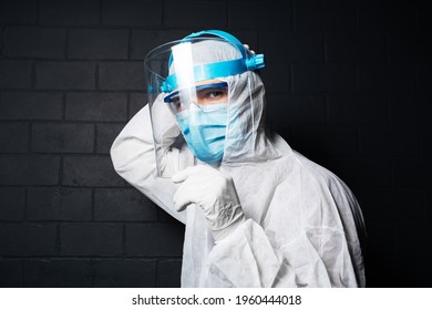 Close-up Portrait Of Young Doctor Man Wearing PPE Suit Against Coronavirus And Covid-19. Background Of Black Brick Wall. Holding Safety Face Shield With Hands.