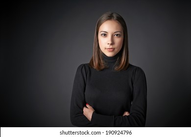 Closeup Portrait Of Young Female In Black Pullover With  Folded Hands With A Speculative Look On Her Face Looking At The Camera, Over Dark Studio Background