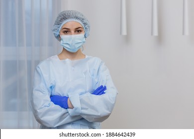 Close-up Portrait A Young Female Doctor In A Protective Medical Mask And Hat. Hispanic Woman Looking At The Camera On A  White Background In A Hospital Room. Portrait Of  Ambulance Nurse