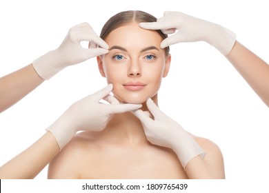 Close-up portrait of young female standing with naked skin, face is touched by doctors in gloves, preparing her for plastic surgery procedures, isolated on white background