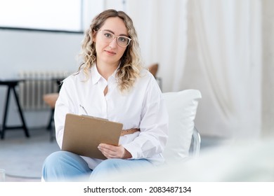 Close-up Portrait Of A Young Female Therapist. Large Glasses For Vision, Psychological Help. Wavy Blonde Hair. There Is A Large Light Window In The Background.