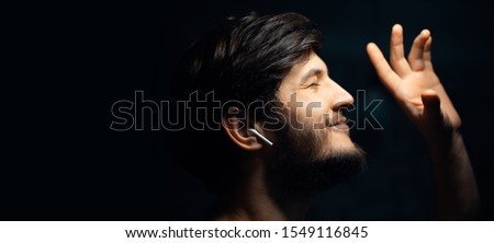 Close-up portrait of young hipster man, listen the music with wireless earphones, on black background.