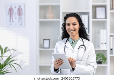 Close-up portrait of a young Latin American female doctor standing in the office in a white coat, holding a tablet and smiling at the camera. - Powered by Shutterstock