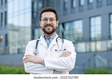 Close-up portrait of a young male doctor in a white coat and glasses standing outside a medical building, arms crossed over his chest and smiling confidently at the camera. - Powered by Shutterstock