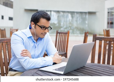 Closeup Portrait, Young Man In Blue Shirt And Black Glasses, Listening To Headphones, Browsing Digital Computer Laptop, Isolated Background Of Sunny Outdoor, Gray Building With Brown Chairs Background