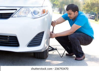 Closeup Portrait, Young Man In Blue Shirt And Black Jeans Happily Fixing Flat Tire With Jack And Tire Iron, Isolated Green Trees And Road Outside Background. Roadside Assistance Concept