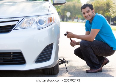 Closeup Portrait, Young Man In Blue Shirt And Black Jeans Happily Fixing Flat Tire With Jack And Tire Iron, Isolated Green Trees And Road Outside Background. Roadside Assistance Concept