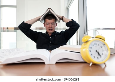 Closeup portrait of young man surrounded by tons of books, alarm clock, stressed from project deadline, study, exams. Negative emotion facial expression feelings, body language