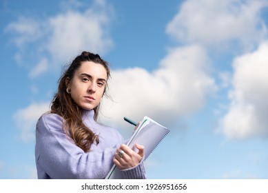 Closeup Portrait Of A Young Pretty Woman Writting On A Notebook While Looking With Serious Face To The Camera With A Sky Full Of Clouds As Background.