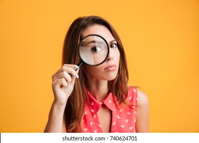 Close-up portrait of young serious woman looking through a magnifying glass, isolated over yellow background