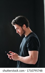 Close-up portrait of a young stylish man with a beard and dark hair in a black T-shirt. A man stands in profile and uses a smartphone on a black background.