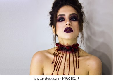 Close-up portrait of young stylish sexy brunette girl with dark bloody makeup with blood drops which is standing and looking straight on white background, halloween concept, free space on left side