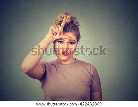 Closeup portrait young unhappy woman giving loser sign on forehead, looking at you, disgust on face isolated on gray wall background. Negative human emotion facial expression