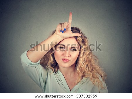 Closeup portrait young unhappy woman giving loser sign on forehead, looking at you, disgust on face isolated on gray wall background. Negative human emotion facial expression