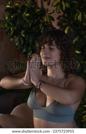 Close-up portrait of a young woman with curly dark hair meditating in a low light setting with her hands in prayer pose