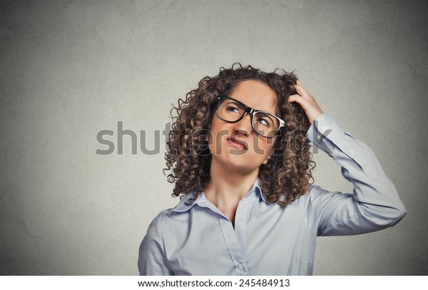 Closeup portrait young woman with glasses\
scratching head, thinking daydreaming something looking up isolated\
grey wall background. Human facial expression emotion feeling body\
language perception