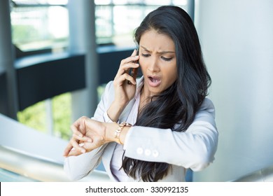 Closeup portrait, young woman in gray business suit blazer talking on cell phone concerned about running out of time on watch, isolated indoors office background