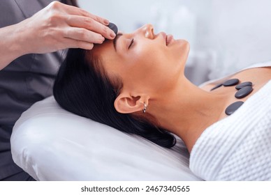 Close-up of a portrait of a young woman with her eyes closed receiving a facial massage with hot stones. A beautiful girl lies and relaxes during a spa procedure. Taking care of yourself concert.