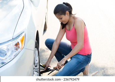 Closeup Portrait, Young Woman In Pink Tanktop And Blue Jeans Fixing Flat Tire With Jack And Tire Iron, Isolated Green Trees And Road Outside Background. Roadside Assistance Concept