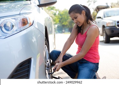 Closeup Portrait, Young Woman In Pink Tanktop And Blue Jeans Fixing Flat Tire With Jack And Tire Iron, Isolated Green Trees And Road Outside Background. Roadside Assistance Concept