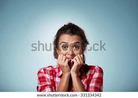 Close-up portrait of a young woman scared ,afraid and anxious biting her finger nails, looking at camera with wide opened eyes isolated on a blue background. Human emotions