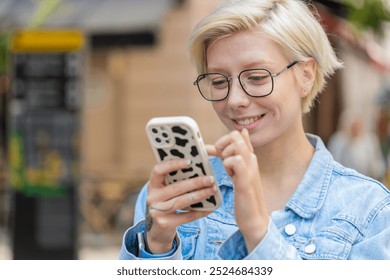 Closeup portrait young woman using smartphone typing texting social media messages e-mail looking for a way on map in mobile navigator app outdoors. Smiling traveler in glasses standing on city street - Powered by Shutterstock