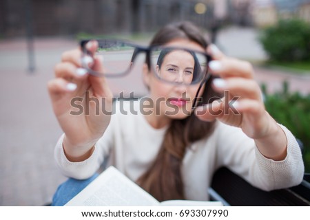 Closeup portrait of young women with glasses. She has eyesight problems and is squinting his eyes a little bit. Beautiful girl is holding eyeglasses right in front of camera with two hand.