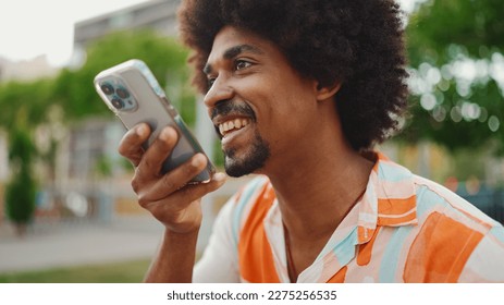 Close-up porttrait of young African American man wearing shirt sitting on park bench using his smartphone. Smiling man sends voice message on mobile phone. - Powered by Shutterstock