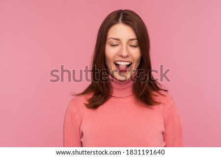 Closeup positive naughty woman with brown hair in pink sweater showing tongue standing with closed eyes, bragging, having fun, childish behaviour. Indoor studio shot isolated on pink background