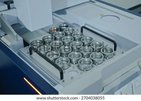 A close-up of a precision autosampler tray inside a laboratory analysis instrument, holding multiple stainless steel crucibles or sample cups. Ideal for automated chemical testing and quality control.