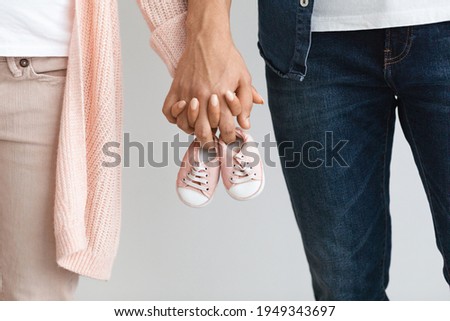 Closeup of pregnant couple carrying small baby shoes while holding hands together, unrecognizable loving man and woman expecting baby, enjoying future parenthood, cropped image, free space