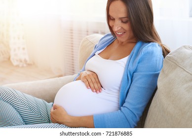 Closeup of pregnant woman holding her belly with arms while sitting on sofa  - Shutterstock ID 721876885
