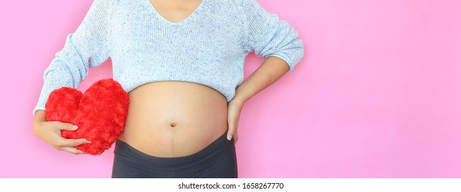 Close-up pregnant woman holding red heart pillow at belly isolated on pink background with copy space.
